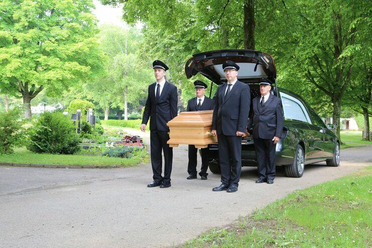 Vier Trauergäste in Anzügen tragen einen Holzsarg neben einem schwarzen Leichenwagen auf einem Friedhof.