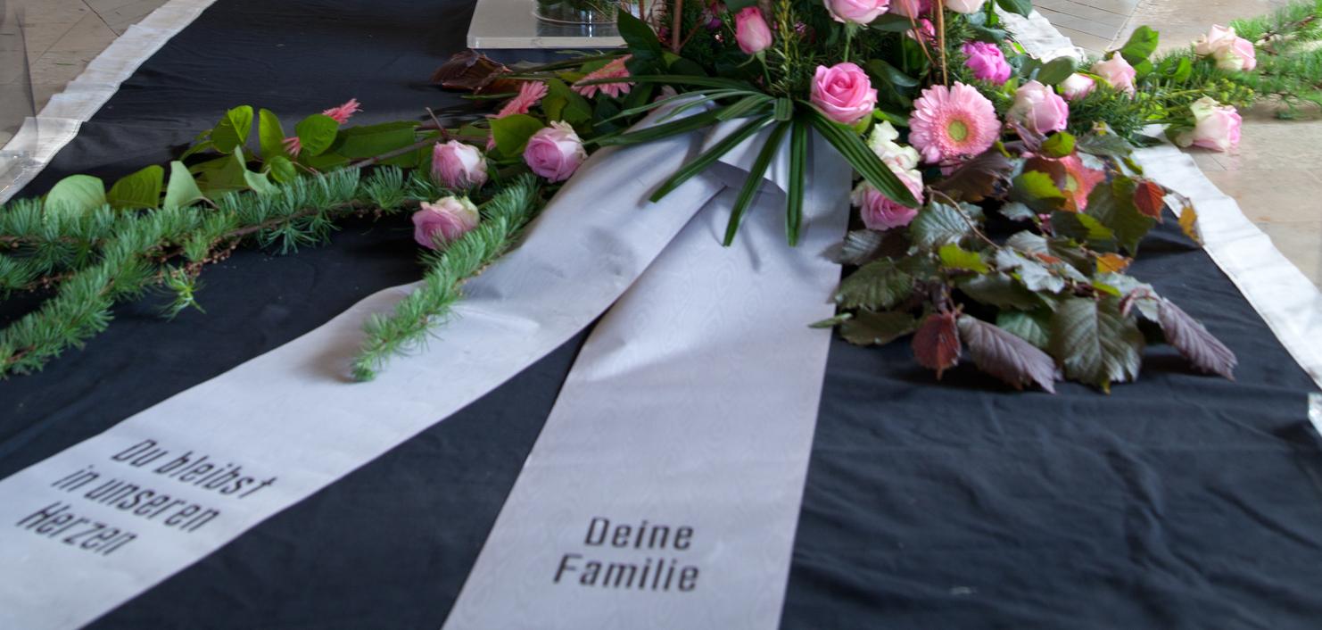Blumenarrangement mit rosa Rosen und eine Trauerschleife vor einem Kreuz in einer Kapelle.