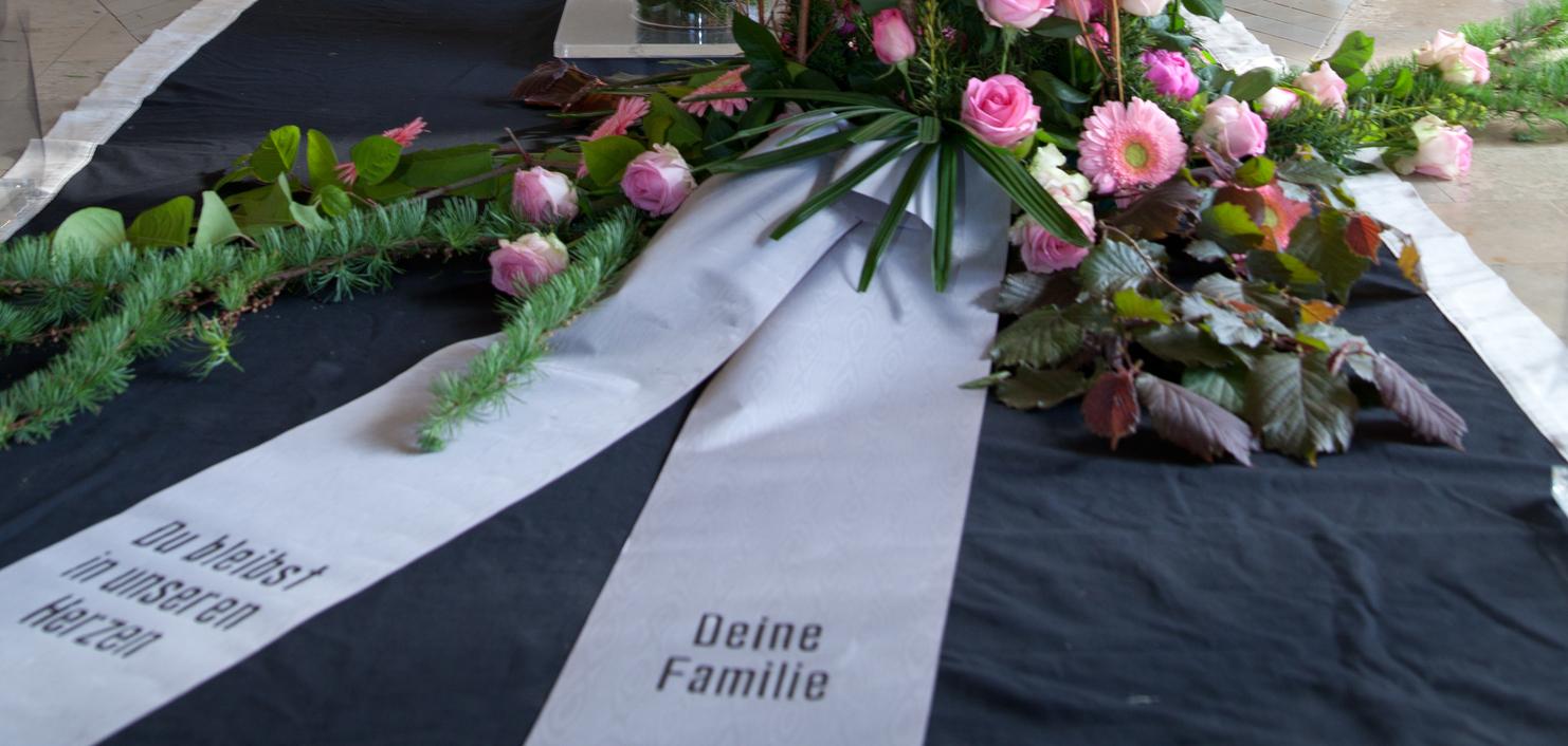 Blumenarrangement mit rosa Rosen und eine Trauerschleife vor einem Kreuz in einer Kapelle.