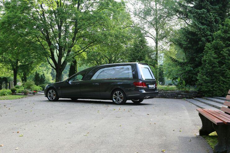 Dunkler Leichenwagen parkt auf einem Weg zwischen Bäumen auf einem Friedhof, neben einer Bank.