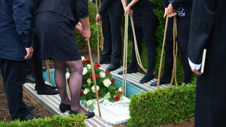 Teilnehmer einer Trauerfeier halten Seile, um Blumenschmuck auf einen Sarg in einer Grabstelle abzusenken.