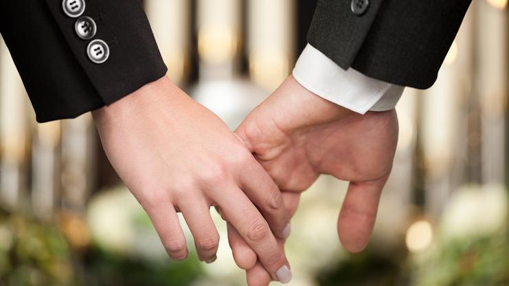 Hände eines Paares halten sich, im Hintergrund verschwommene Kerzen und florale Dekorationen.