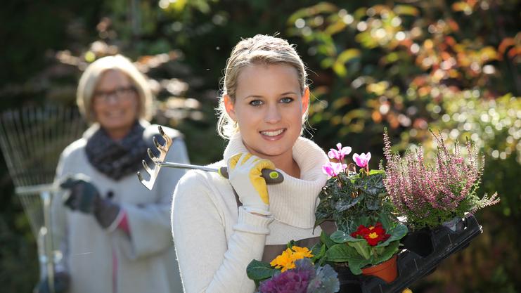 Frau mit Pflanzen in Töpfen und einer Gartengabel lächelt, während eine zweite Person im Hintergrund mit einer Harke steht.