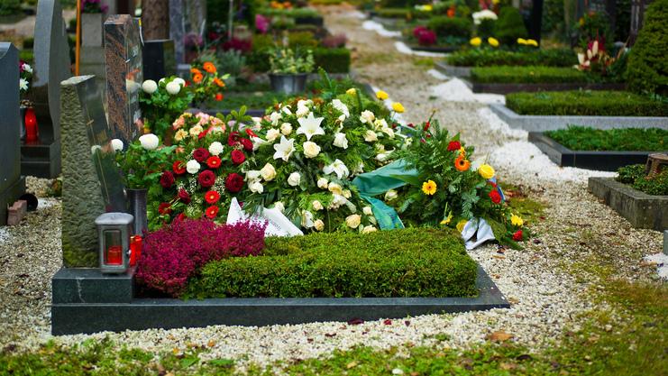 Blühende Grabstätte mit zahlreichen bunten Blumenarrangements und einer trauernden Person im Hintergrund.