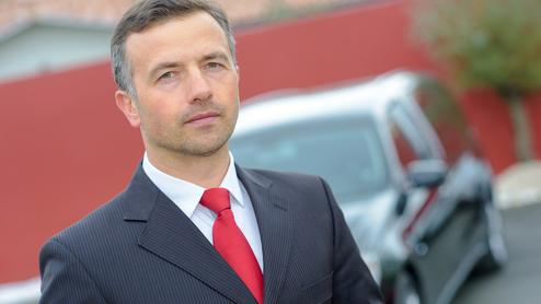 Mann in Anzug mit roter Krawatte steht vor einem schwarzen Auto und einem roten Hintergrund.