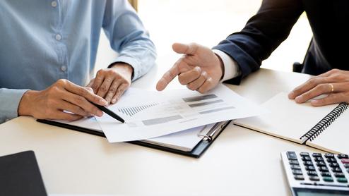 Zwei Personen diskutieren über Dokumente mit Finanzgraphen an einem Bürotisch, Notizblock und Taschenrechner liegen bereit.