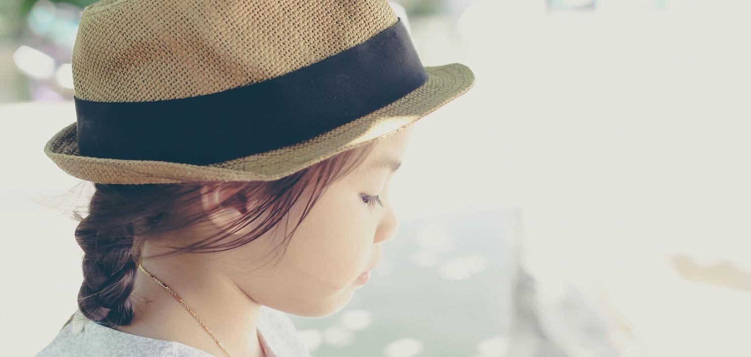 Portrait eines Mädchens mit Zopf und Sonnenhut, das nachdenklich zur Seite schaut. Heller Hintergrund mit verschwommenen Farben.