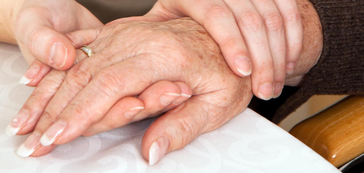 Zwei Hände, eine ältere und eine jüngere, liegen auf einem Tisch, wobei die jüngere Hand die ältere sanft umschließt.