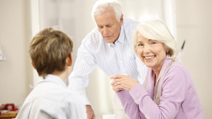 Lächelnde ältere Frau sitzt am Tisch, während ein Mann im Hintergrund Getränke serviert, ein Junge schaut sie an.